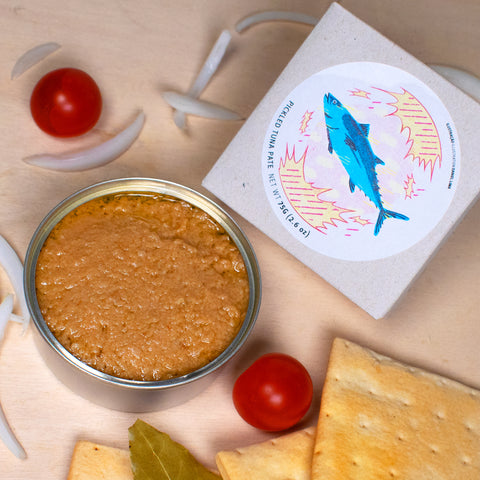 Conserve de pâté de thon ouverte sur une table en bois clair entouré de tomates cerises oignons et cracottes