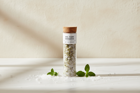 tube de sel avec bouchon en liège sur une table en marbre blanc avec graines de sel et feuilles d'origan sur fond blanc texturé