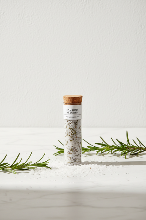 tube de sel et romarin avec bouchon en liège sur une table en marbre blanc entouré de grains de sel et feuilles de romarin sur fond blanc texturé